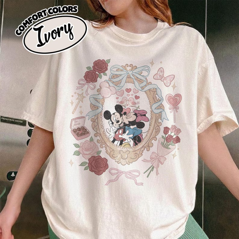 Puede incluir: Una camiseta de color marfil con un gr&aacute;fico de Mickey y Minnie Mouse bes&aacute;ndose dentro de un marco ovalado ornamentado. El dise&ntilde;o incluye rosas rojas, una cinta azul, una caja de bombones con la etiqueta Be Mine, una piruleta y un ramo.