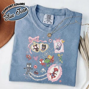 K&ouml;nnte beinhalten: Blaues Jeans-T-Shirt mit einer Grafik verschiedener Cartoon-Superhelden. Das Shirt hat einen Rundhalsausschnitt und kurze &Auml;rmel. Die Grafik zeigt Spider-Man, Black Panther und andere Charaktere. Das Shirt ist aus weichem Material.