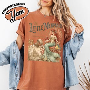 以下が含まれることがあります： 錆色のTシャツ。ヴィンテージスタイルのリトルマーメイドのイラストが特徴です。デザインには、貝殻に座るアリエル、タツノオトシゴ、馬車、城が含まれています。シャツには「Comfort Colors」と「Yam」のロゴが入っています。