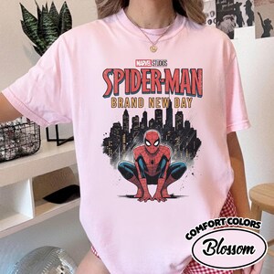Puede incluir: Una camiseta de manga corta de color rosa claro con un estampado gr&aacute;fico de Spider-Man agachado frente a un horizonte urbano negro. El texto dice Marvel Studios Spider-Man Brand New Day. La camiseta tiene cuello redondo y un dise&ntilde;o holgado.