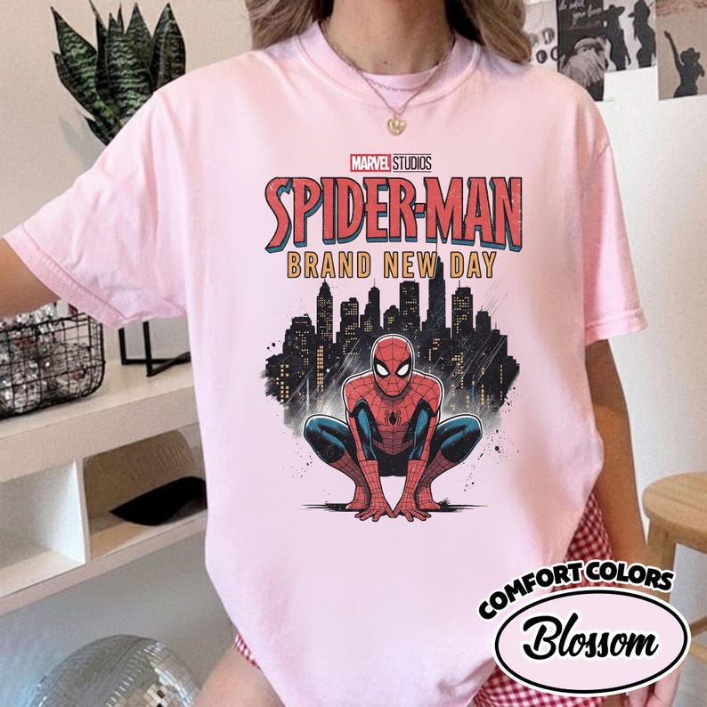 Puede incluir: Una camiseta de manga corta de color rosa claro con un estampado gr&aacute;fico de Spider-Man agachado frente a un horizonte urbano negro. El texto dice Marvel Studios Spider-Man Brand New Day. La camiseta tiene cuello redondo y un dise&ntilde;o holgado.