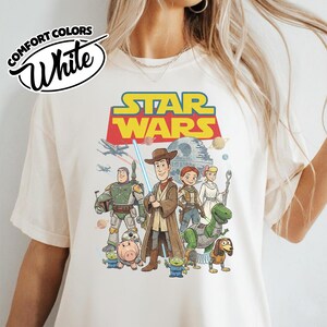 May include: White Comfort Colors t-shirt featuring a Star Wars graphic with characters from Toy Story. The design includes Woody, Buzz Lightyear, and other characters with the Star Wars logo in yellow and red.