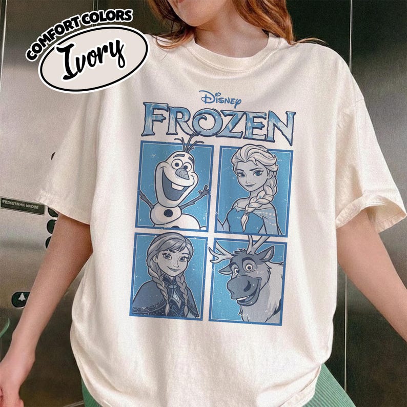 May include: Ivory-colored t-shirt featuring the Disney Frozen logo and character illustrations. The shirt displays Olaf, Elsa, Anna, and Sven in blue-framed squares. The words "COMFORT COLORS" and "Ivory" are printed on the top left.
