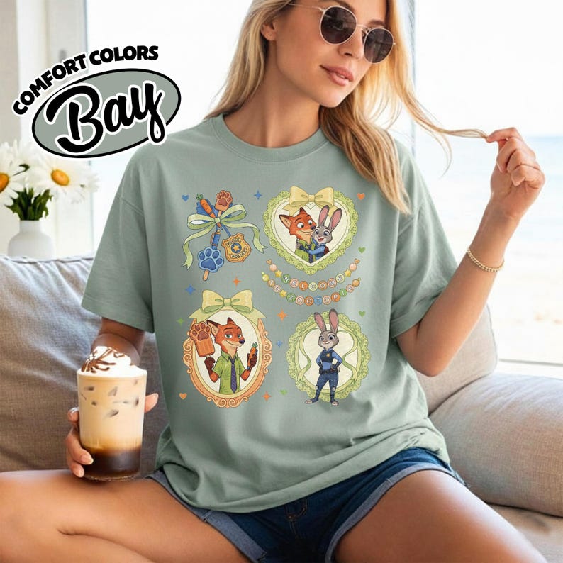 May include: A sage green short-sleeve t-shirt featuring a graphic print of characters from Zootopia. The design includes four framed illustrations of a fox and a rabbit, a police badge, a paw print, and a carrot, all in pastel colors.