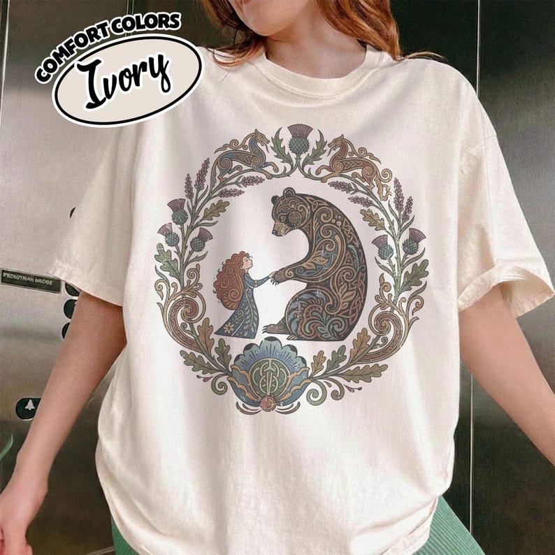 May include: Ivory-colored t-shirt featuring a circular design with a bear and a woman, surrounded by floral and animal motifs. The shirt has short sleeves and a crew neck. The text "COMFORT COLORS" and "Ivory" are printed on the top left.
