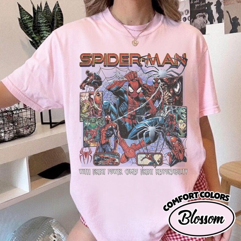 K&ouml;nnte beinhalten: Hellrosa T-Shirt mit einem Spider-Man-Motiv. Das Design beinhaltet das Wort "SPIDER-MAN" in einer Retro-Schriftart sowie Comic-Panels von Spider-Man in Aktion. Der Text "WITH GREAT POWER COMES GREAT RESPONSIBILITY" ist ebenfalls sichtbar.