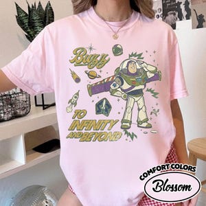 May include: A light pink short-sleeve t-shirt featuring a graphic of the character Buzz Lightyear saluting. The design includes space-themed elements like a rocket, planet, and comet, with the text "Buzz TO INFINITY AND BEYOND" in yellow.