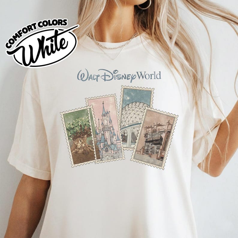 Op de afbeelding: Cr&egrave;mekleurig T-shirt met de tekst "Walt Disney World" boven vier postzegelachtige illustraties. De postzegels tonen verschillende Disney park bezienswaardigheden. Het shirt heeft het "Comfort Colors" en "White" logo linksboven.
