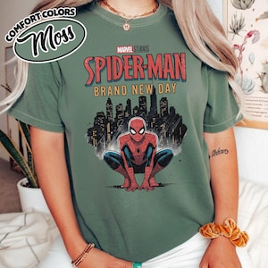 Puede incluir: Una camiseta de manga corta color verde salvia con un gr&aacute;fico de Spider-Man agachado frente a un horizonte urbano negro. El texto dice Marvel Studios Spider-Man Brand New Day. La camiseta tiene cuello redondo y un dise&ntilde;o de corte holgado.