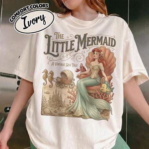 以下が含まれることがあります： 「リトル・マーメイド」のヴィンテージイラストが描かれたアイボリー色のTシャツ。デザインには、貝殻の中にいるアリエル、フランダー、セバスチャン、お城などが含まれています。Tシャツには「Comfort Colors」と「Ivory」の文字が入っています。