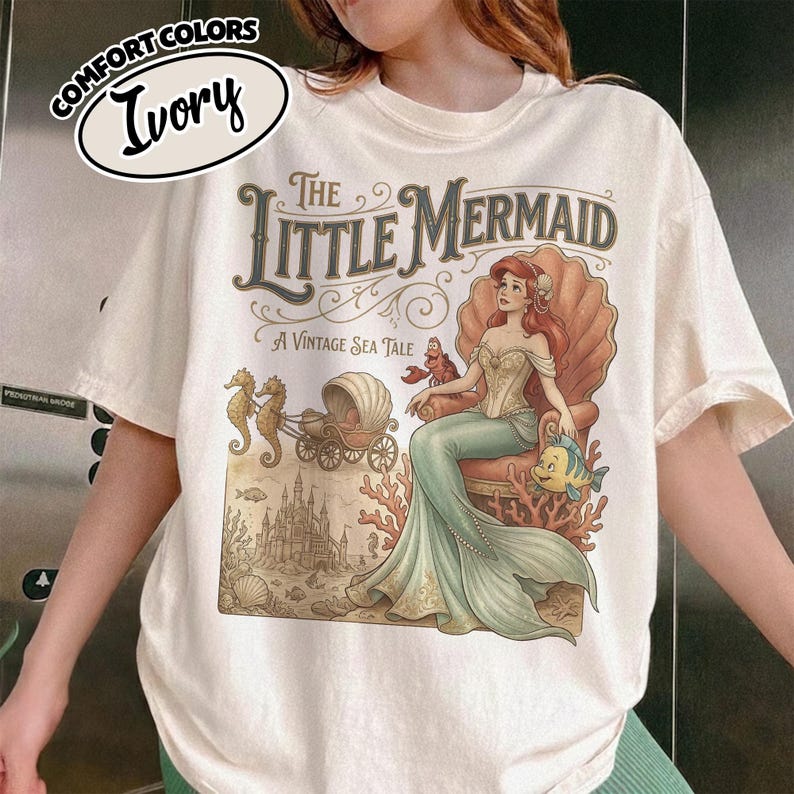 以下が含まれることがあります： 「リトル・マーメイド」のヴィンテージイラストが描かれたアイボリー色のTシャツ。デザインには、貝殻の中にいるアリエル、フランダー、セバスチャン、お城などが含まれています。Tシャツには「Comfort Colors」と「Ivory」の文字が入っています。