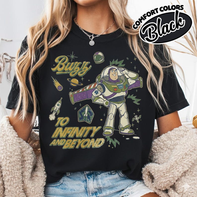 May include: A black t-shirt featuring a graphic print of Buzz Lightyear saluting, surrounded by space-themed elements including a rocket, a planet, and stars. The text reads Buzz TO INFINITY AND BEYOND in a distressed yellow font style.