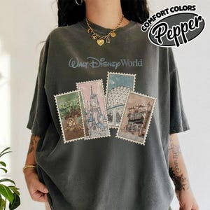 Op de afbeelding: Donkergrijs T-shirt met de tekst "Walt Disney World" boven vier postzegelachtige afbeeldingen van Disney parkattracties. Het shirt heeft de woorden "Comfort Colors" en "Pepper" in een cirkelvormig ontwerp.