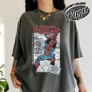 K&ouml;nnte beinhalten: Dunkelgraues T-Shirt mit einem Comic-Grafik von Spider-Man. Das Design enth&auml;lt den Text "the AMAZING SPIDER-MAN" und "PETER PARKER ALIAS SPIDER-MAN". Das T-Shirt hat kurze &Auml;rmel und eine lockere Passform.