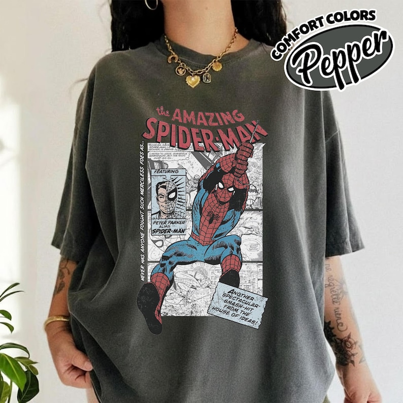 K&ouml;nnte beinhalten: Dunkelgraues T-Shirt mit einem Comic-Grafik von Spider-Man. Das Design enth&auml;lt den Text "the AMAZING SPIDER-MAN" und "PETER PARKER ALIAS SPIDER-MAN". Das T-Shirt hat kurze &Auml;rmel und eine lockere Passform.