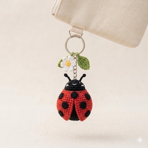 May include: A crochet ladybug keychain featuring a red body with black spots, a black head with two antennae, and a small white flower with a yellow center and a green leaf attached to the silver metal ring, hanging from a beige fabric pouch.
