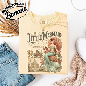 以下が含まれることがあります： 淡い黄色のComfort Colors Tシャツ。リトルマーメイドのヴィンテージイラストが特徴です。イラストは、アリエルが貝殻の玉座に座り、タツノオトシゴ、馬車、城が描かれています。シャツには「Comfort Colors」と「Banana」の文字が入っています。