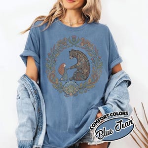 May include: Blue t-shirt with a circular design featuring a bear and a girl holding hands, surrounded by floral and leaf elements. The bear has intricate patterns. The shirt has the words "COMFORT COLORS" and "Blue Jean" in a circular design.