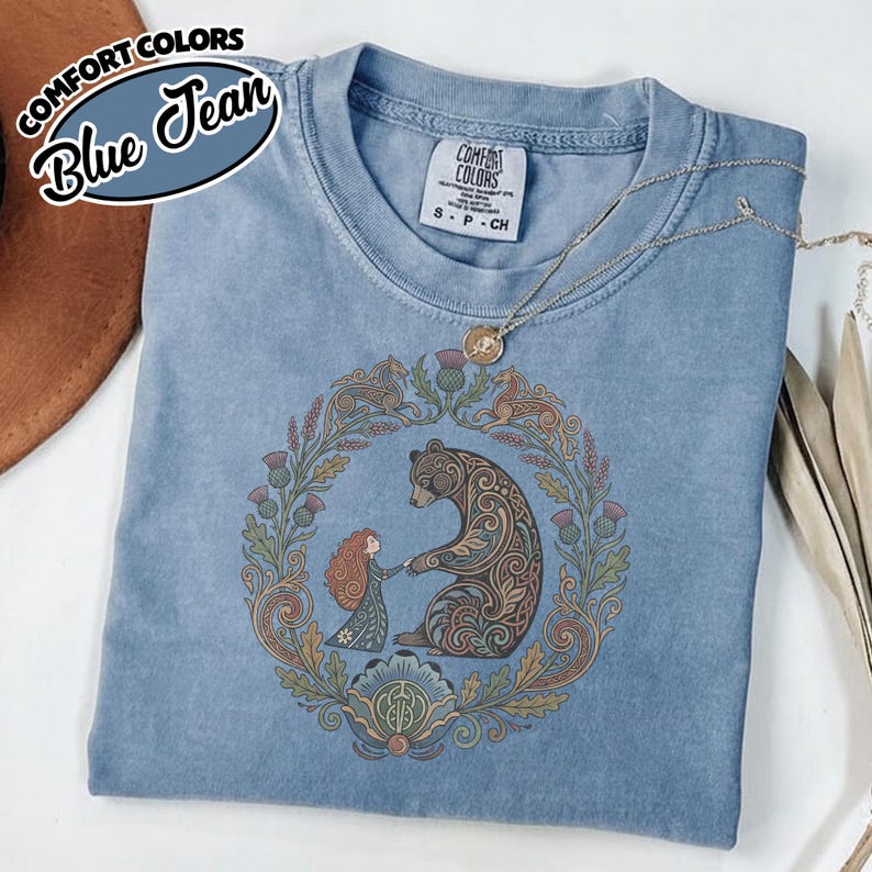 May include: Blue jean Comfort Colors t-shirt featuring a circular design with a bear and a girl holding hands, surrounded by a floral wreath. The shirt has a small Comfort Colors tag and a necklace. The text "Comfort Colors Blue Jean" is in the upper left corner.