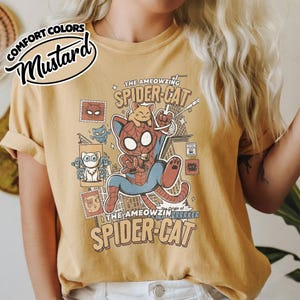May include: Mustard-colored t-shirt featuring a cartoon illustration of a cat dressed as Spider-Man, with the text "THE AMEOWZING SPIDER-CAT". The design includes comic-style elements and the phrase "Origin of Brrrker".