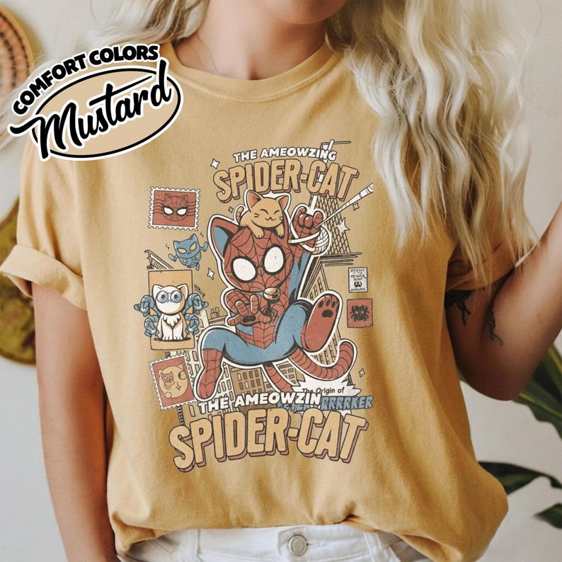 May include: Mustard-colored t-shirt featuring a cartoon illustration of a cat dressed as Spider-Man, with the text "THE AMEOWZING SPIDER-CAT". The design includes comic-style elements and the phrase "Origin of Brrrker".