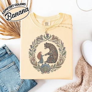 May include: A light yellow Comfort Colors t-shirt with a circular design featuring a bear and a girl holding hands, surrounded by a floral wreath. The shirt has the text "Comfort Colors Banana" in a black and white oval.