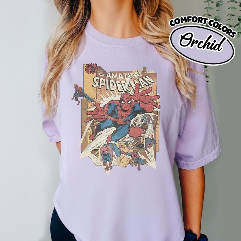 K&ouml;nnte beinhalten: Ein helllila T-Shirt mit einem Vintage-Comic-Design von Spider-Man. Das Design enth&auml;lt den Text "The Amazing Spider-Man" und die Worte "Comfort Colors Orchid" in einem kreisf&ouml;rmigen Logo.