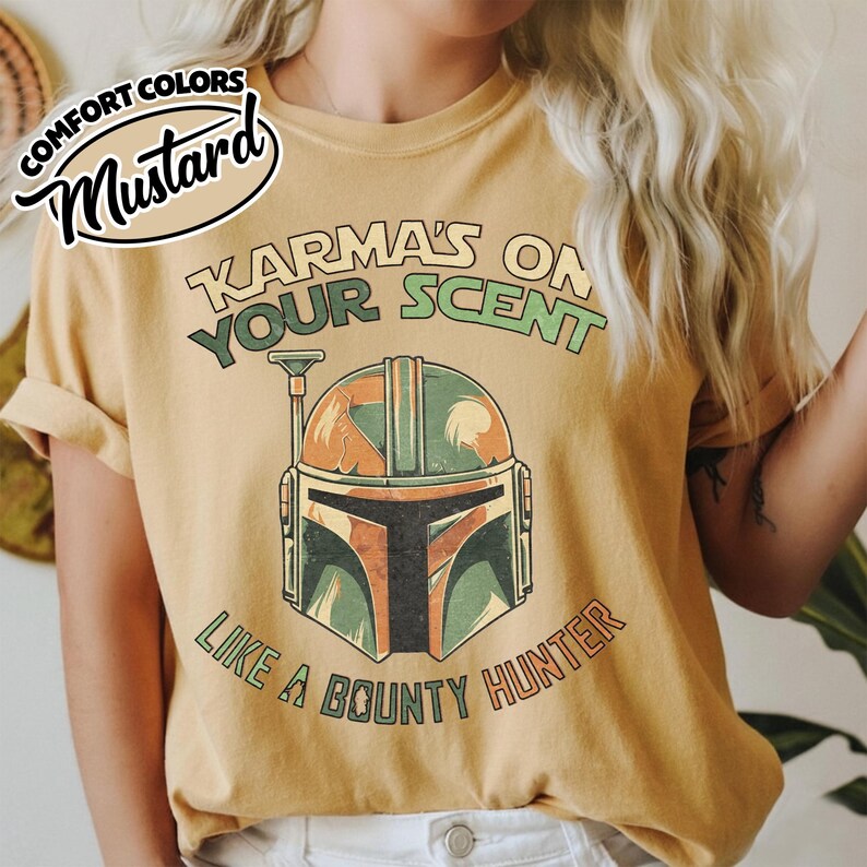 May include: Mustard-colored t-shirt with the text "Karma's on Your Scent Like a Bounty Hunter" and a graphic of a Mandalorian helmet in green, orange, and brown. The shirt has rolled-up sleeves and a relaxed fit.