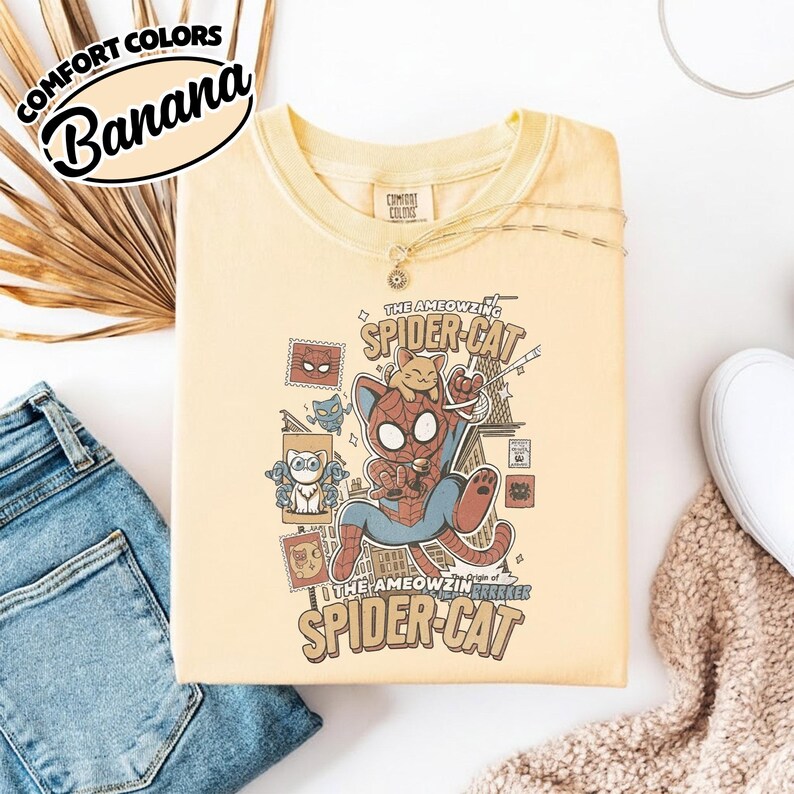 May include: A light yellow Comfort Colors t-shirt with a graphic of a cat dressed as Spider-Man, with the text "The AMEOWZING SPIDER-CAT". The shirt also features the word "Banana" in a black oval.