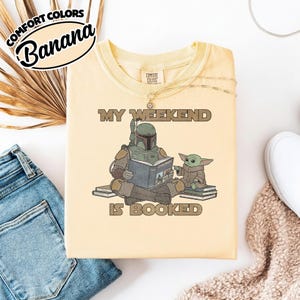 Może przedstawiać: Jasnoż&oacute;łta koszulka z napisem "MY WEEKEND IS BOOKED" i grafiką Boba Fetta czytającego książkę z Grogu. Koszulka ma etykietę Comfort Colors w lewym g&oacute;rnym rogu.