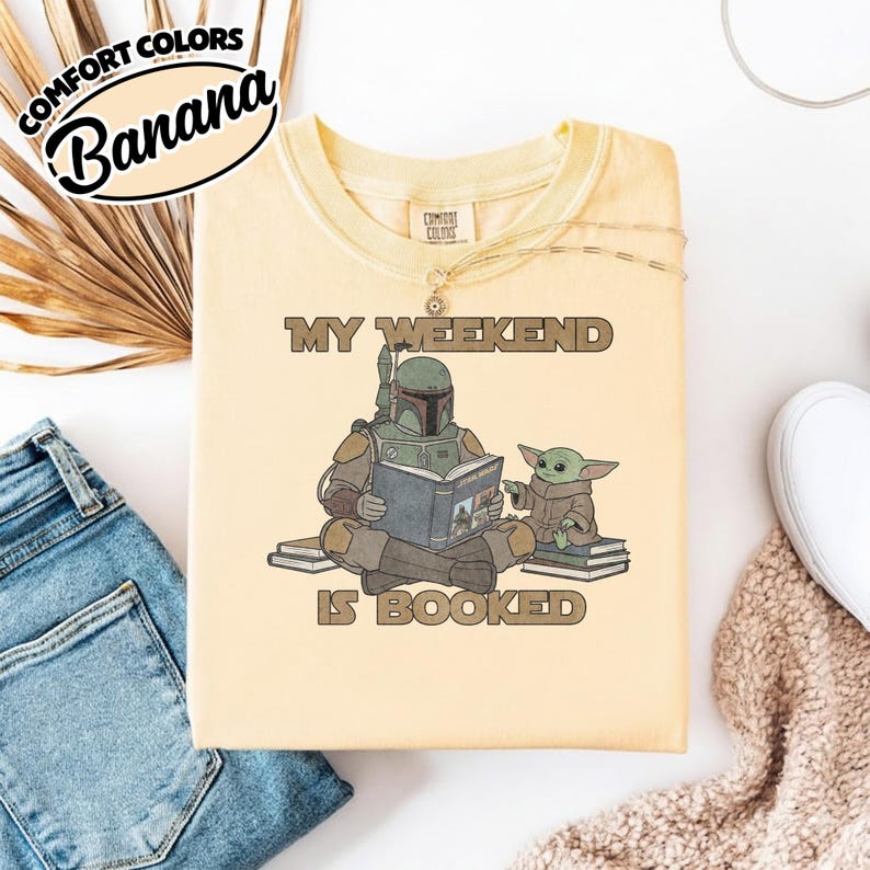 Może przedstawiać: Jasnoż&oacute;łta koszulka z napisem "MY WEEKEND IS BOOKED" i grafiką Boba Fetta czytającego książkę z Grogu. Koszulka ma etykietę Comfort Colors w lewym g&oacute;rnym rogu.