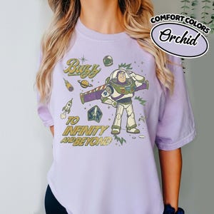 May include: A light purple t-shirt featuring a graphic of the character Buzz Lightyear saluting. The design includes space-themed elements like a rocket, planet, and stars, with the text "Buzz TO INFINITY AND BEYOND" printed in a distressed yellow font.