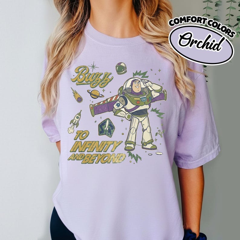 May include: A light purple t-shirt featuring a graphic of the character Buzz Lightyear saluting. The design includes space-themed elements like a rocket, planet, and stars, with the text "Buzz TO INFINITY AND BEYOND" printed in a distressed yellow font.