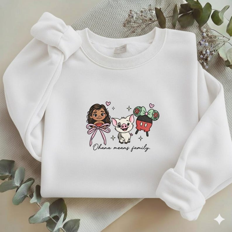 May include: A white crewneck sweatshirt featuring a central embroidered graphic of a cartoon character with long brown hair, a small pig, and a pair of mouse ears. The text "Ohana means family." is embroidered in black script below the characters.