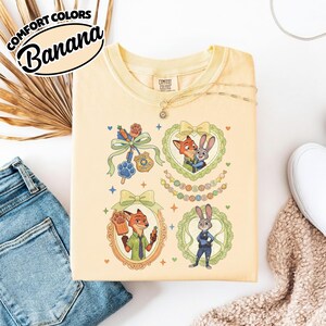 May include: A pale yellow t-shirt featuring a graphic print of characters from Zootopia, including Nick Wilde and Judy Hopps, framed by decorative bows and borders, with a small police badge and paw print icon, and the text Welcome to Zootopia.