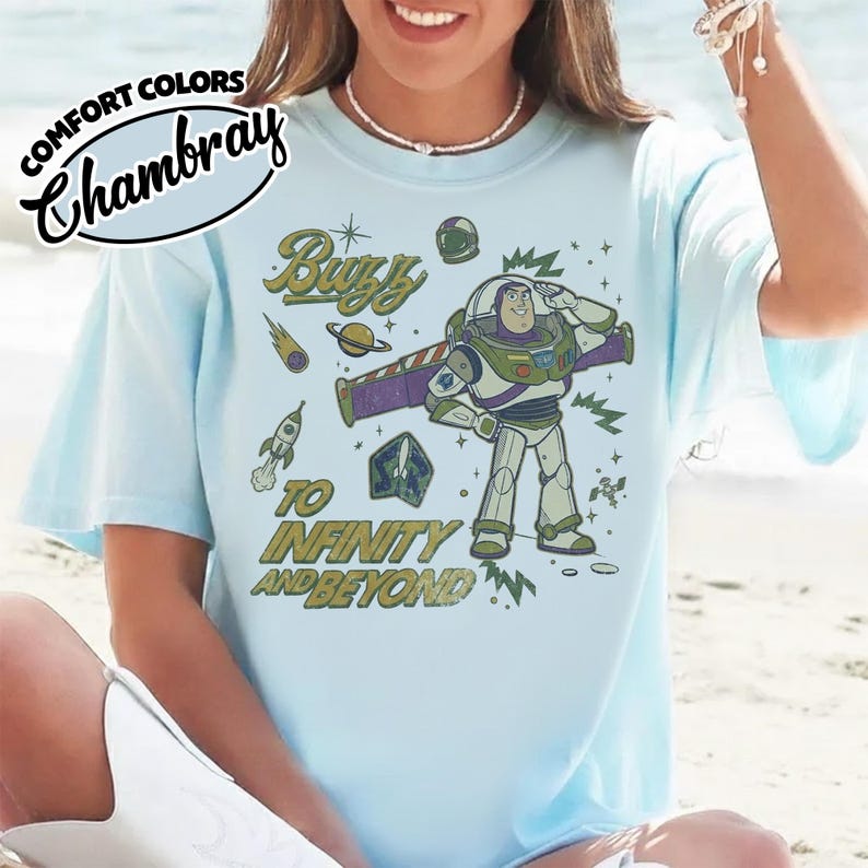 May include: A light blue t-shirt featuring a graphic of Buzz Lightyear saluting, surrounded by space-themed icons including a rocket, planet, and stars. The text reads "Buzz" and "To Infinity and Beyond" in a distressed green font. A "Comfort Colors Chambray" logo is visible in the corner.