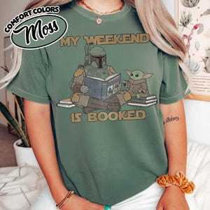 Może przedstawiać: Zielona koszulka z napisem "MY WEEKEND IS BOOKED" i grafiką Boba Fetta czytającego książkę z Grogu. Koszulka ma luźny kr&oacute;j i jest wykonana z miękkiego materiału. Koszulka ma r&oacute;wnież słowa "COMFORT COLORS" i "Moss" w okręgu.