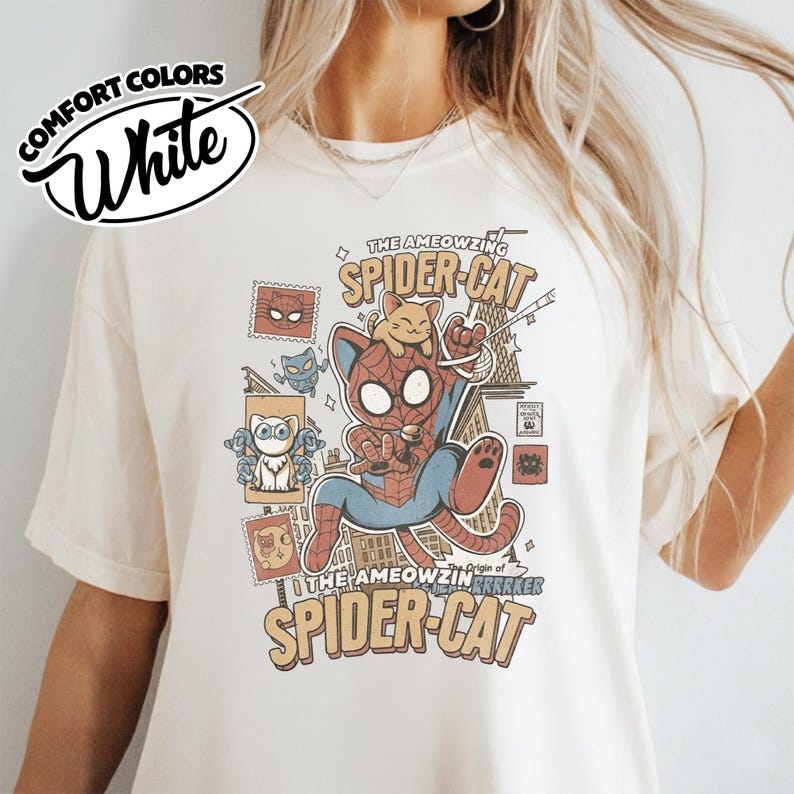 May include: White Comfort Colors t-shirt featuring a comic-style illustration of a cat dressed as Spider-Man, with the text "THE AMEOWZING SPIDER-CAT" and "Origin of the AMEOWZIN GRARKER". The design includes various cat-themed comic book elements.