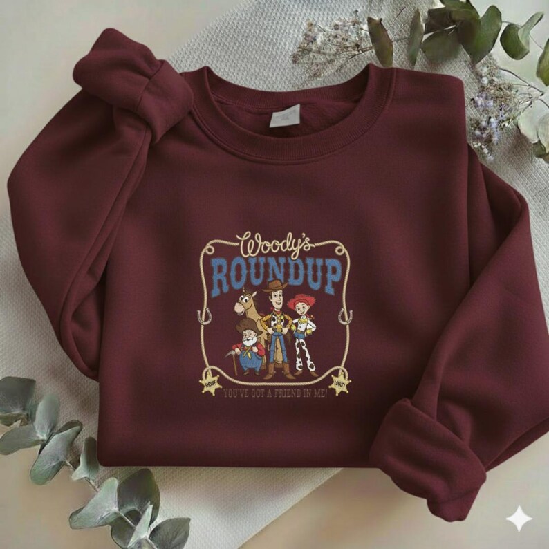 Puede incluir: Una sudadera de cuello redondo color granate con un estampado gr&aacute;fico de Woody, Jessie, Bullseye y Stinky Pete de Toy Story. El texto dice Woody's ROUNDUP y YOU'VE GOT A FRIEND IN ME, enmarcado por un dise&ntilde;o de lazo con dos estrellas de sheriff.