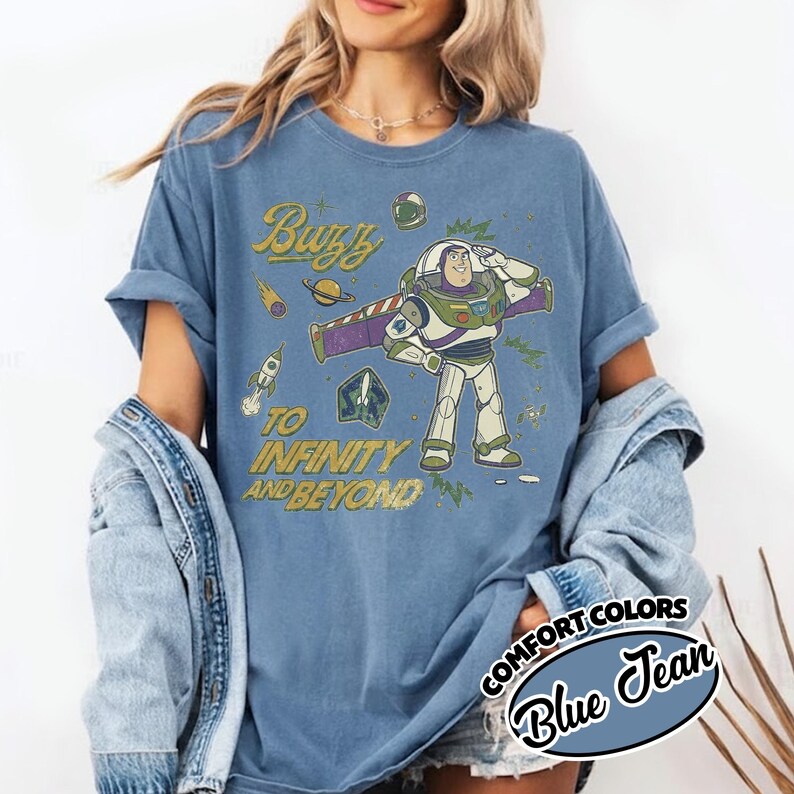 May include: A blue jean colored t-shirt featuring a graphic of the character Buzz Lightyear. The design includes the text "Buzz" and "To Infinity and Beyond" in a yellow font, surrounded by illustrations of a rocket, a planet, and stars.
