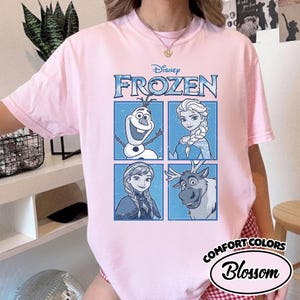 May include: A light pink t-shirt featuring the Disney movie "Frozen." The design includes the word "Frozen" in blue, along with images of Olaf, Elsa, Anna, and Sven. The shirt also has the "Comfort Colors" and "Blossom" logo.