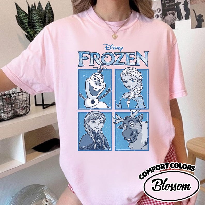 May include: A light pink t-shirt featuring the Disney movie "Frozen." The design includes the word "Frozen" in blue, along with images of Olaf, Elsa, Anna, and Sven. The shirt also has the "Comfort Colors" and "Blossom" logo.