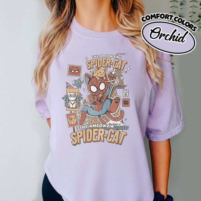 May include: Orchid-colored t-shirt featuring a cartoon illustration of a cat dressed as Spider-Man, with the text "THE AMEOWZING SPIDER-CAT" and "COMFORT COLORS" above. The design includes a small cat on top of the Spider-Cat.