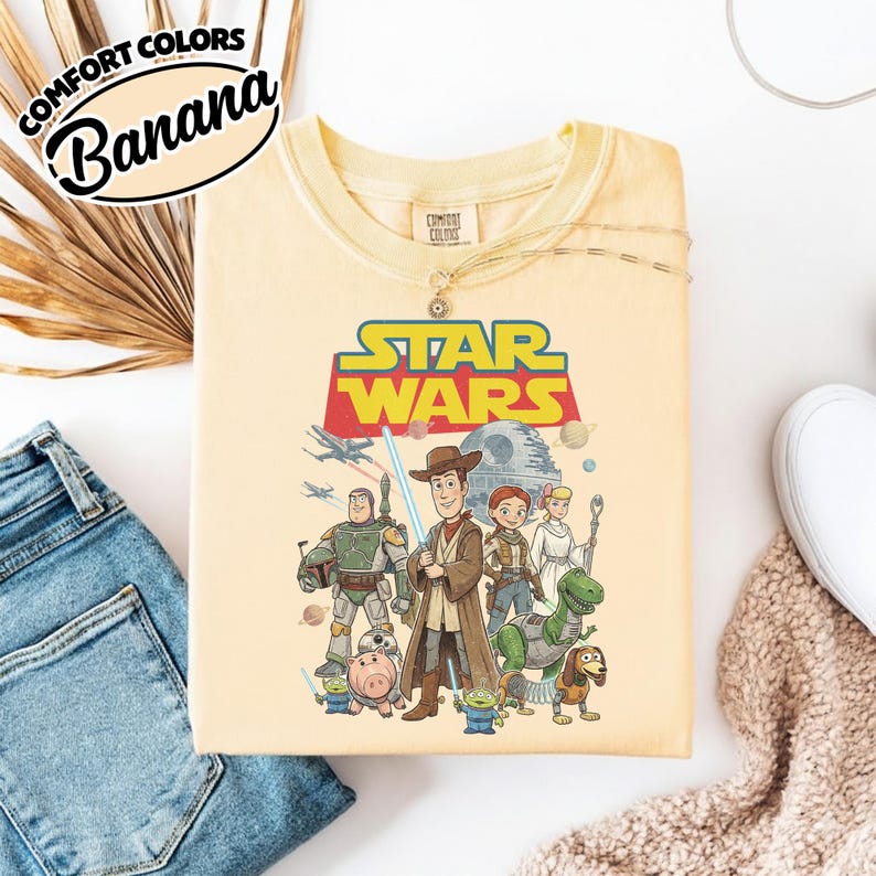 May include: A light yellow t-shirt featuring a Star Wars graphic with characters from the movie and Toy Story. The shirt has the words "COMFORT COLORS" and "Banana" printed on it.