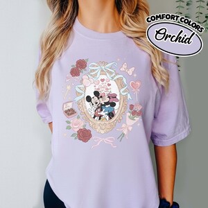 Puede incluir: Una camiseta de color morado claro con un gr&aacute;fico central de Mickey y Minnie Mouse dentro de un marco ovalado ornamentado. El dise&ntilde;o incluye rosas, una caja de bombones, una cinta azul y corazones peque&ntilde;os. El texto Comfort Colors Orchid es visible.