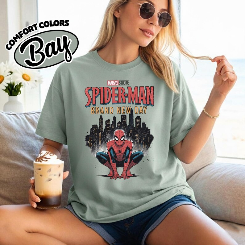 Puede incluir: Una camiseta de manga corta en color verde salvia con un gr&aacute;fico de Spider-Man agachado frente a un horizonte urbano negro. El texto dice Marvel Studios Spider-Man Brand New Day. La camiseta tiene cuello redondo y un dise&ntilde;o holgado.