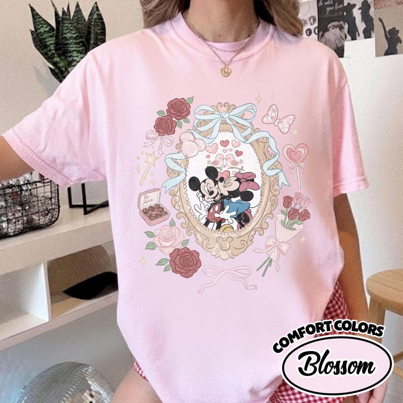Puede incluir: Una camiseta rosa claro con un gr&aacute;fico central de Mickey y Minnie Mouse bes&aacute;ndose dentro de un marco dorado ornamentado. El dise&ntilde;o incluye cintas azul claro, rosas rojas y rosas, una caja de bombones con la etiqueta Be Mine y un ramo de rosas.