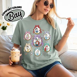 May include: Sage green t-shirt featuring framed portraits of animated princesses. The portraits are surrounded by decorative bows and mirrors. The shirt has the text "COMFORT COLORS Bay" in a circular design.