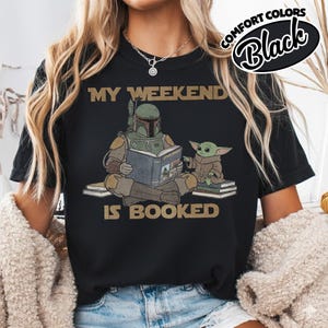 Może przedstawiać: Czarny t-shirt ze złotym napisem "MY WEEKEND IS BOOKED". Grafika przedstawia postać w hełmie czytającą książkę z małą zieloną istotą. Słowa "COMFORT COLORS Black" są w czarnym owalu.