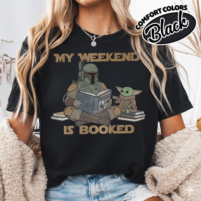 Może przedstawiać: Czarny t-shirt ze złotym napisem "MY WEEKEND IS BOOKED". Grafika przedstawia postać w hełmie czytającą książkę z małą zieloną istotą. Słowa "COMFORT COLORS Black" są w czarnym owalu.
